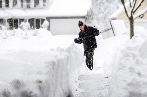 A person shoveling snow from a walkway, surrounded by deep snow and a snowy landscape.
