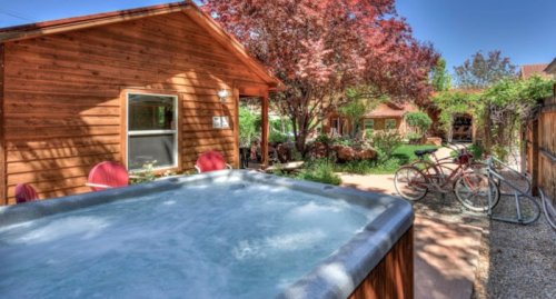 A hot tub in a backyard with a wooden cabin, colorful trees, and bicycles nearby on a sunny day.