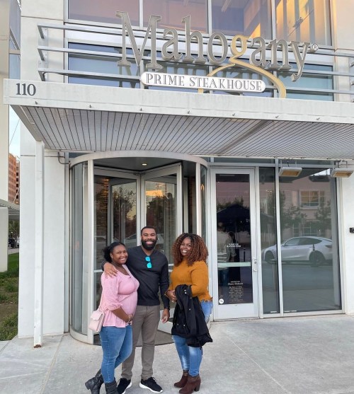 Three friends pose outside Mahogany Prime Steakhouse, smiling in front of the entrance.