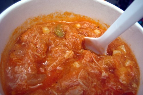 A close-up of a bowl of thick, orange soup with shredded chicken and vegetables, accompanied by a white spoon.