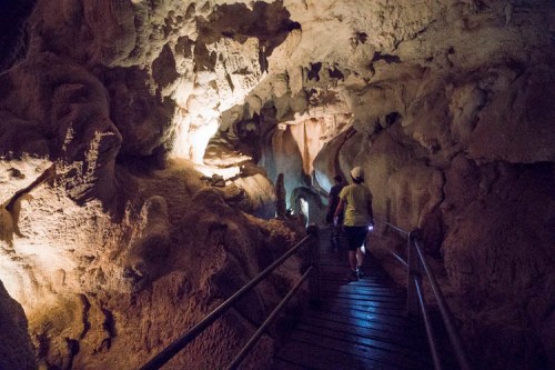 A person walks along a wooden path through a dimly lit cave with textured rock formations.