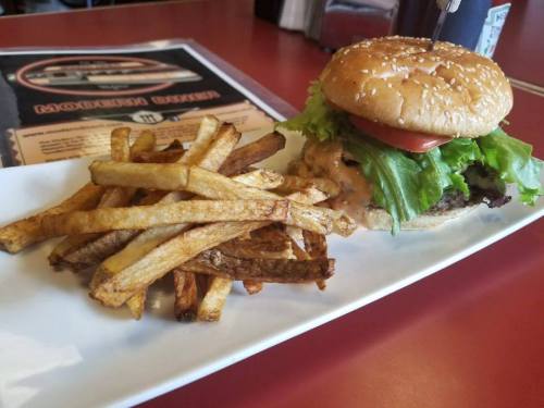 A sesame seed bun burger with lettuce, tomato, and sauce, served with a side of golden French fries on a white plate.