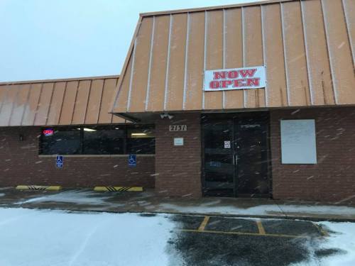 A storefront with a "NOW OPEN" sign, snow falling, and a clear view of the entrance and parking area.
