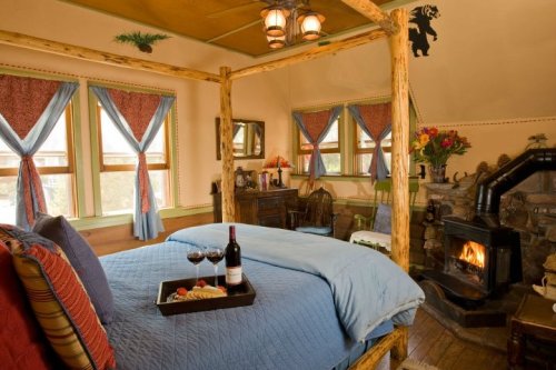 Cozy bedroom with a four-poster bed, fireplace, and a tray of wine and snacks, surrounded by rustic decor.