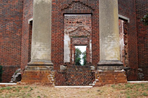 Ruins of a brick building with tall columns and multiple doorways, surrounded by grass and trees.