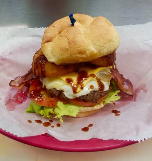 A stacked burger with bacon, onion rings, lettuce, tomato, and sauce, served on a bun with a toothpick on top.