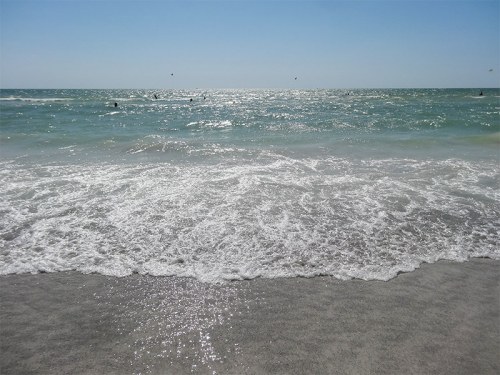 Waves gently lap at a sandy beach under a clear blue sky, with distant figures enjoying the water.