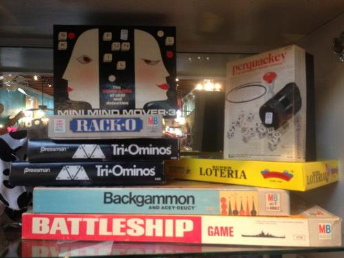 A collection of board games and puzzles stacked on a shelf, including Battleship, Backgammon, and Tri-Ominos.