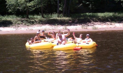 A group of people on yellow inflatable rafts, smiling and waving, enjoying a sunny day on a river.