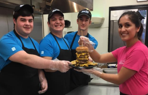 Four smiling restaurant staff members pose with a large cheeseburger on a plate, showcasing their teamwork and food.