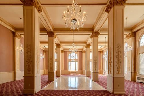 Elegant interior of a spacious room with chandeliers, tall columns, and large windows, featuring a patterned carpet.