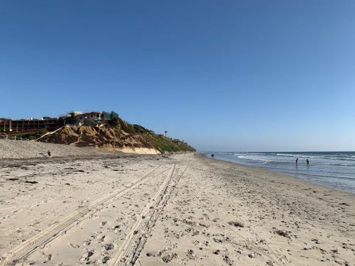 A sandy beach with gentle waves, a cliff in the background, and a clear blue sky. People stroll along the shore.
