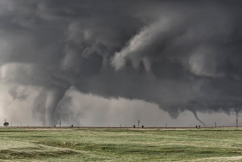 Two tornadoes touch down in a dark, stormy sky over a grassy landscape.