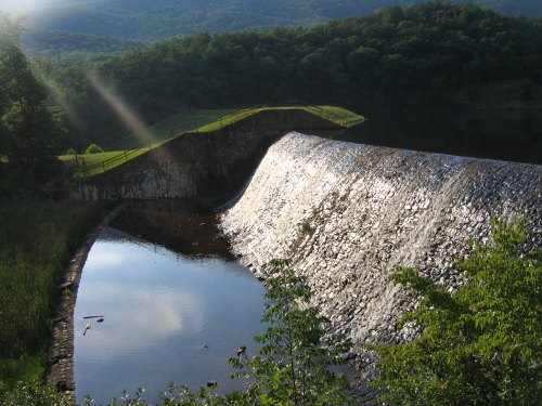 A stone dam spills water into a calm lake, surrounded by lush green hills and trees under a clear sky.