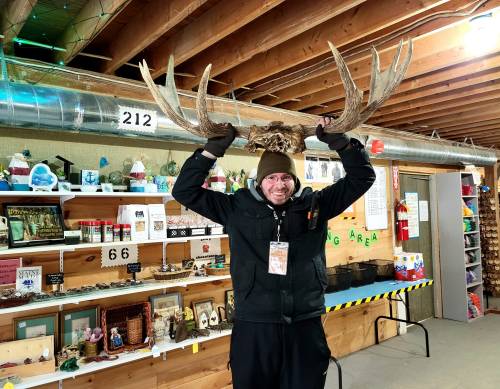 A person in a black jacket and brown hat holds large antlers above their head in a rustic indoor setting.