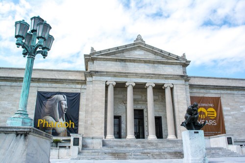 Exterior of a museum featuring columns, banners for an exhibit, and a statue in front.