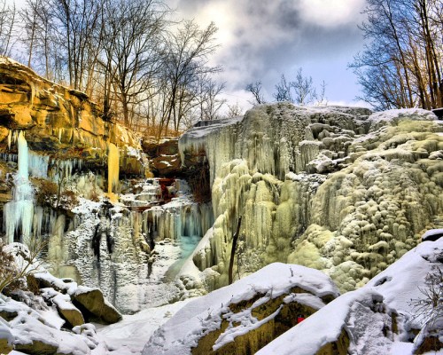 A frozen waterfall surrounded by icy rocks and bare trees under a cloudy sky, creating a serene winter landscape.