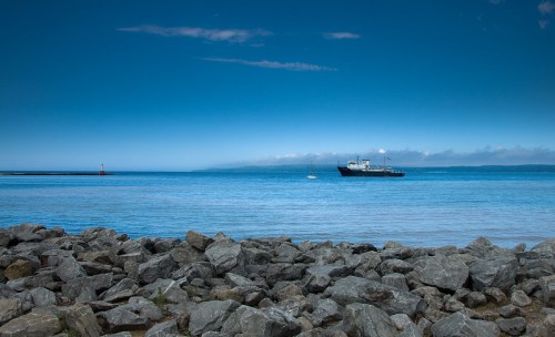 A calm sea with a large ship in the distance, rocky shore in the foreground, and a clear blue sky above.