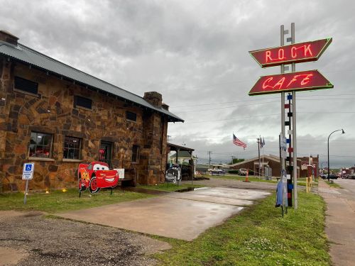 A stone building with a neon "R.O.C.K. Cafe" sign, featuring cartoon car cutouts outside, under a cloudy sky.