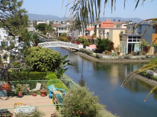 A scenic view of a canal lined with colorful houses, a small bridge, and lush greenery under a clear blue sky.
