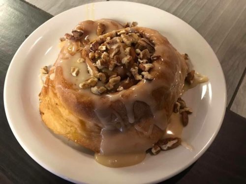 A large cinnamon roll topped with creamy glaze and sprinkled with chopped nuts, served on a white plate.