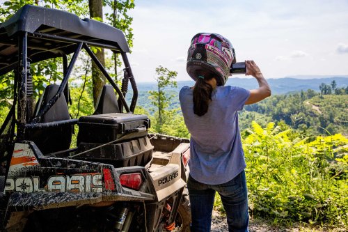 A person in a helmet takes a photo of a scenic view from a forested area, with an ATV parked nearby.