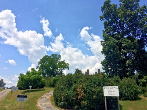 A scenic view of a pathway leading to historic ruins, surrounded by greenery and a bright blue sky with clouds.