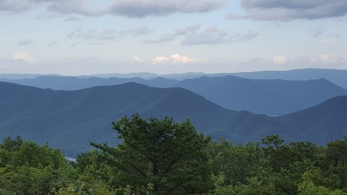 A scenic view of layered blue mountains under a cloudy sky, with green trees in the foreground.