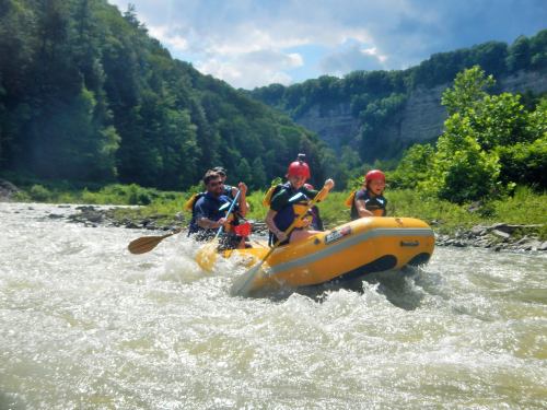 Three people in a yellow raft navigate a river, surrounded by lush green hills and a bright blue sky.