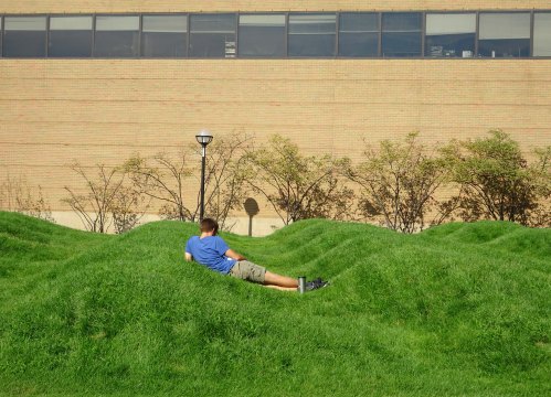 A person relaxes on a grassy, undulating landscape in front of a brick building.