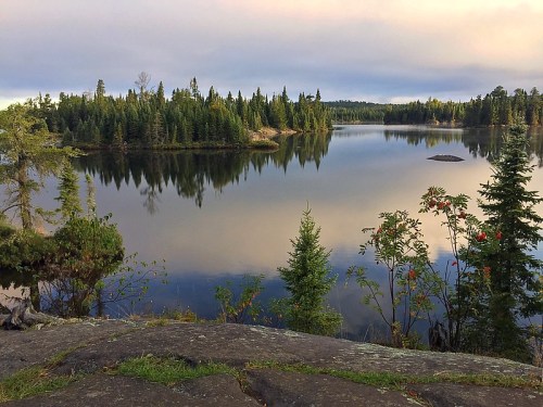 A serene lake surrounded by lush trees, reflecting the sky and landscape, with rocky shores in the foreground.