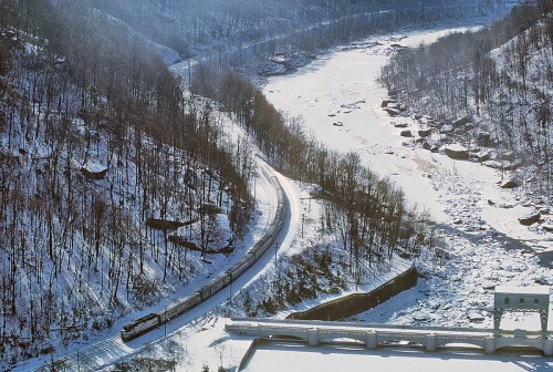 A snowy landscape with a winding train track along a river, surrounded by trees and rocky terrain.