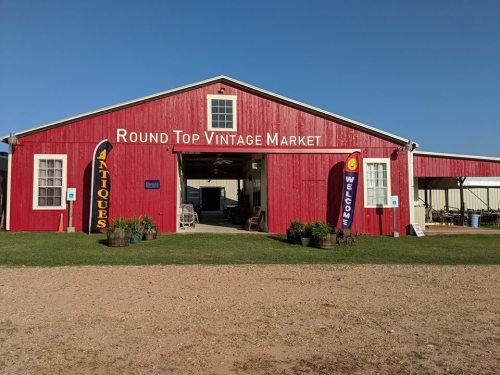 Red barn-style building with "Round Top Vintage Market" sign, featuring antique displays and welcoming banners.