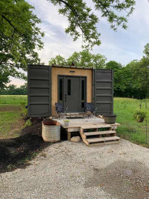 A small, modern cabin made from shipping containers, with a wooden deck and two chairs in a grassy area.