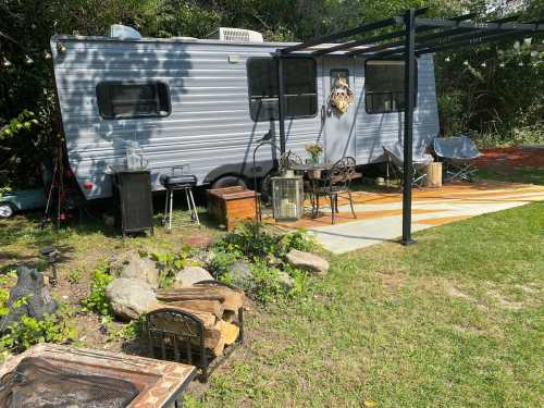A gray camper trailer in a green outdoor setting, with a seating area, fire pit, and decorative plants nearby.
