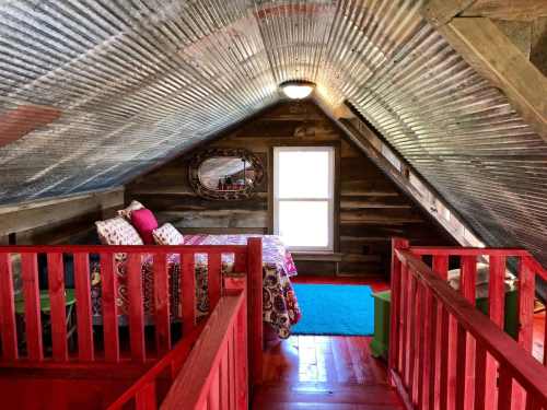 Cozy attic bedroom with a corrugated metal ceiling, red accents, a colorful rug, and a large window.