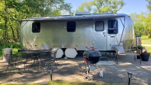 A silver Airstream trailer parked in a wooded area, with a grill and outdoor seating nearby.