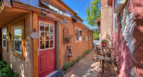 Charming adobe building entrance with a red door, surrounded by greenery and outdoor seating. Bright blue sky above.