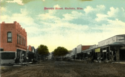Historic postcard of Ennis Street in Shubuta, Mississippi, featuring storefronts and a dirt road under a blue sky.