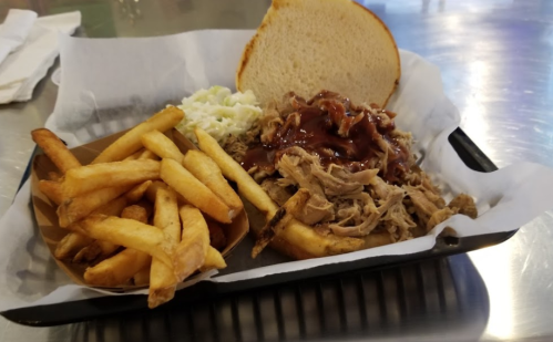 A plate with pulled pork, barbecue sauce, a slice of bread, coleslaw, and a side of crispy French fries.