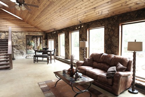 Cozy living room with wooden ceiling, stone walls, a brown sofa, coffee table, and large windows letting in natural light.