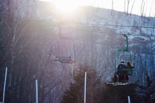 Two people on a ski lift against a backdrop of snowy mountains and a bright sun.