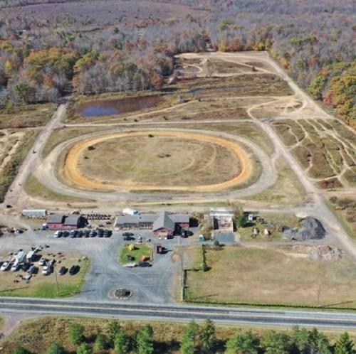 Aerial view of a large, oval dirt track surrounded by trees, with a parking area and buildings nearby.