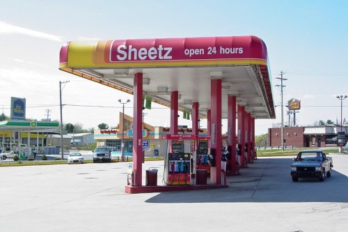 A Sheetz gas station with a red canopy, open 24 hours, surrounded by other businesses and parked cars.
