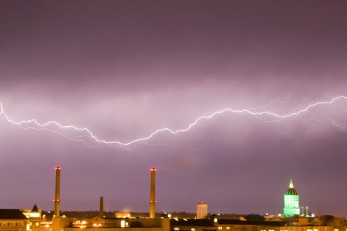 A dramatic night sky illuminated by lightning, with industrial buildings and a green dome in the background.