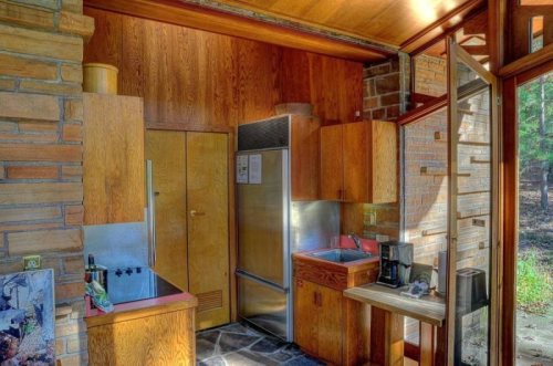 Modern kitchen with wooden cabinetry, stainless steel appliances, and large windows overlooking a natural setting.