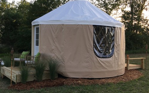 A cozy yurt with a wooden deck, surrounded by greenery, featuring a large window and outdoor seating.