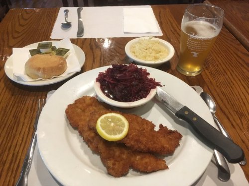 A plate with two fried fish fillets, lemon, red cabbage, and a side of rice, accompanied by a roll and a beer.