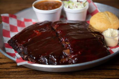 A platter of glazed ribs with sides of baked beans, coleslaw, cornbread, and butter on a checkered tablecloth.