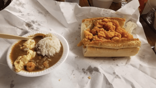 A plate with rice and shrimp stew next to a sandwich filled with fried shrimp on a crumpled paper surface.
